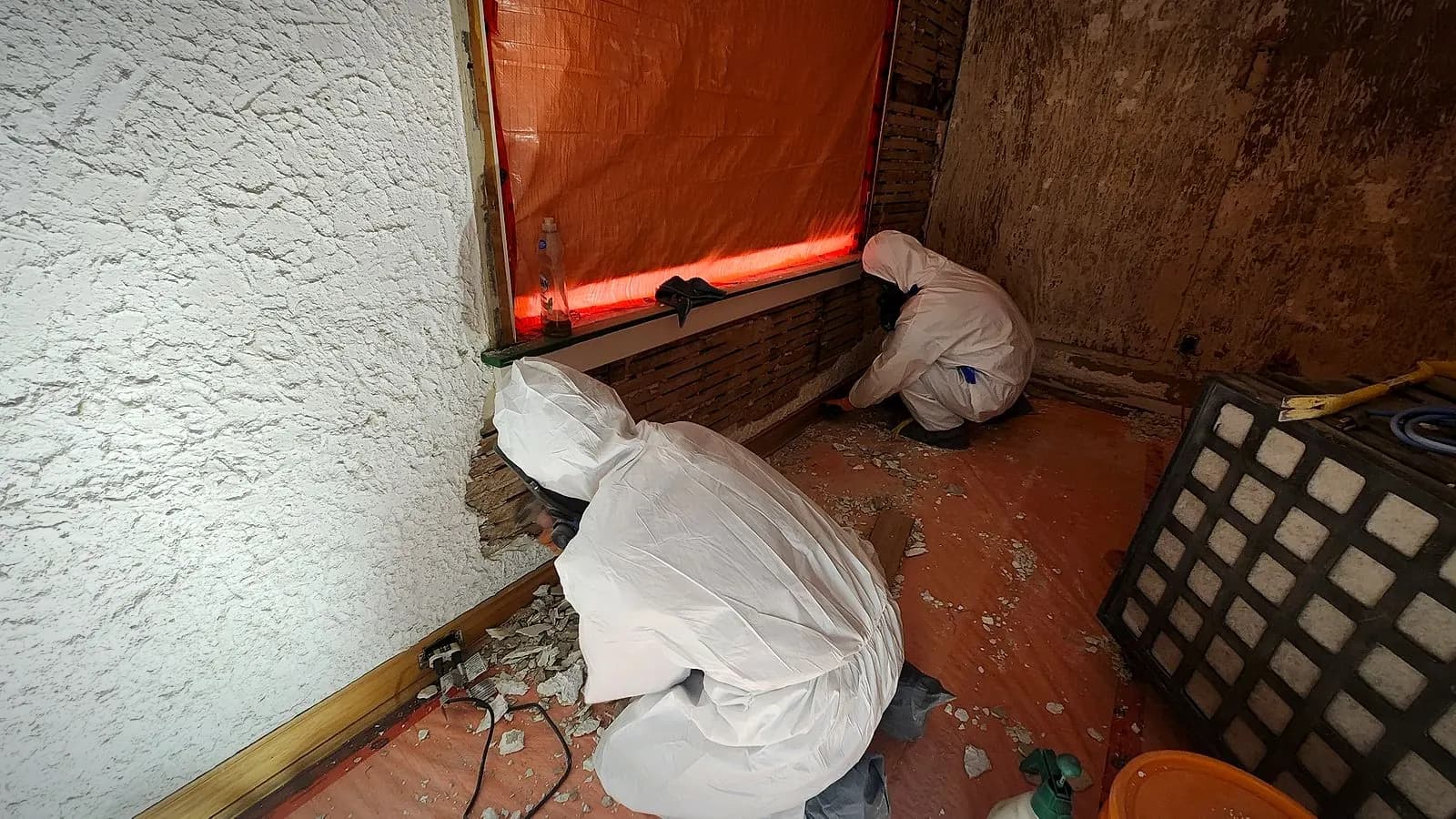 Two Relief Restorations technicians removing asbestos plaster from a wall inside containment in a Winnipeg home