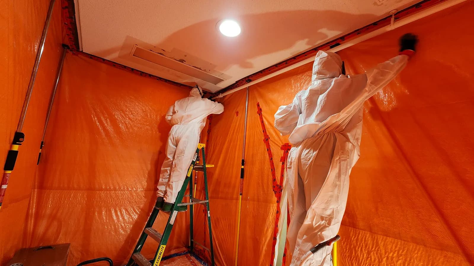 Two Relief Restorations technicians in protective suits removing asbestos drywall inside a sealed containment in a Winnipeg property