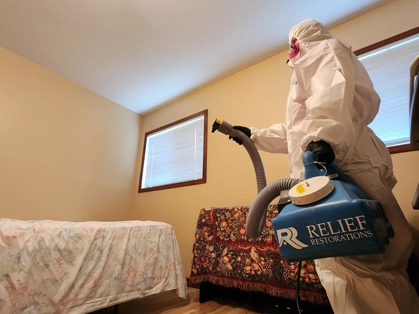 Relief Restorations technician in a full protective suit and respirator operating a ULV fogging machine to disinfect a residential room.